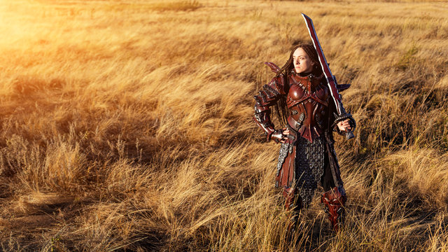 Girl in medieval knight's armor with a big sword against the sunset fields background