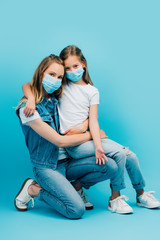 girl in white t-shirt sitting on lap of mother in denim clothes while wearing medical masks and looking at camera on blue