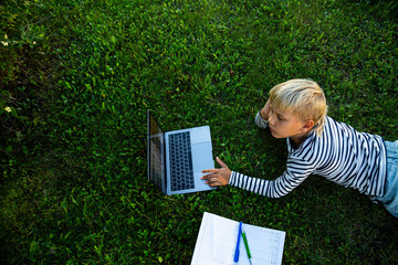 Cute boy lies on the grass with a laptop. Child learning online outdoors.	