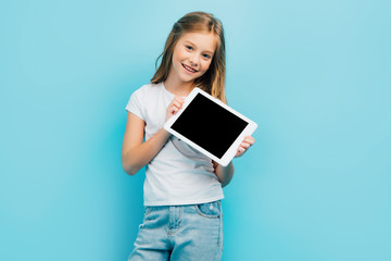 child in white t-shirt and jeans looking at camera while holding digital tablet with blank screen isolated on blue