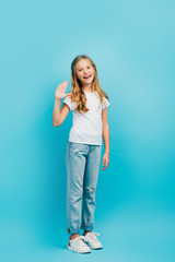 Fototapeta premium full length view of girl in white t-shirt and jeans waving hand while looking at camera on blue