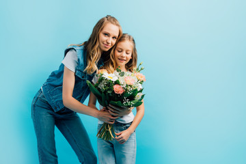 young woman in denim clothes presenting flowers to daughter while looking at camera isolated on blue