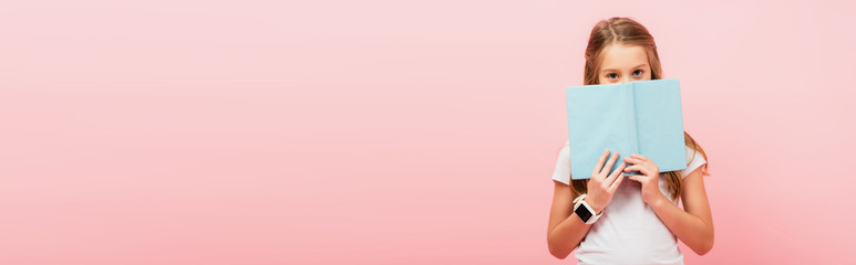 horizontal image of girl in smartwatch obscuring face with book while looking at camera isolated on pink - Powered by Adobe