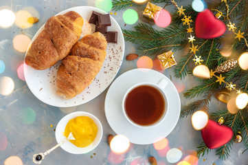 Christmas breakfast with tea and croissants on a blue concrete background with spruce branch and Christmas lights.