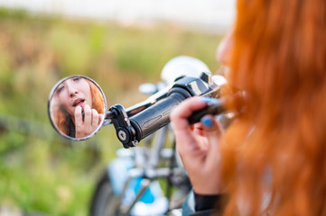 A woman on a motorcycle looks in the mirror and applying lipstick. Reflection of a red-haired biker girl.