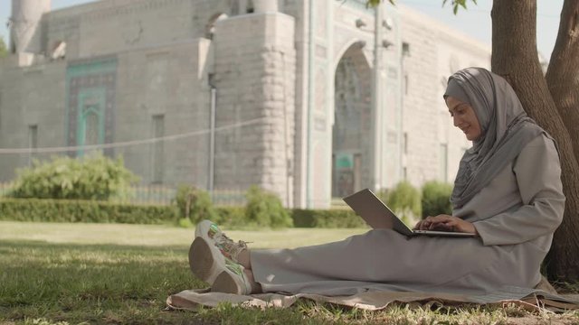 Wide shot of young mixed-race woman wearing hijab and trendy sneakers is sitting on grass leaning on tree trunk near mosque and working on laptop