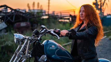 Fototapeta na wymiar Curly red-haired woman in a black leather jacket sits on a motorcycle at sunset. Portrait of a serious girl driving a bike.