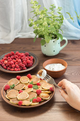 Pancake cereals in your hand on a fork with yogurt. Red ripe fresh raspberries in a brown plate and a bunch of Melissa in a mug on the dark wooden table near the window. Delicious Breakfast