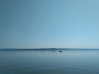 A tiny boat is floating on the blue river, the forest is visible in the distance, the sky is clear and cloudless. On a quiet calm river floats a motor boat. A haze of heat hangs over the river