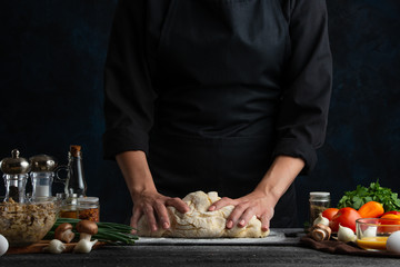 Professional chef works with dough for making pizza, pasta, or bread or pie, against the background of vegetables, culinary recipes and a recipe book