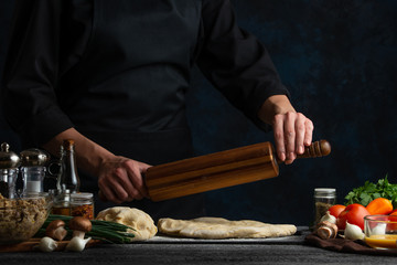 Professional chef works with dough for making pizza, pasta, or bread or pie, against the background of vegetables, culinary recipes and a recipe book