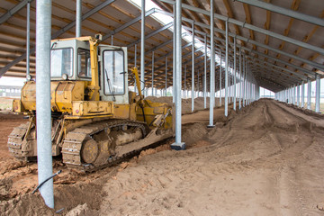 Fototapeta na wymiar A bulldozer is leveling the ground in a barn under construction. Construction of an agricultural complex. Earthworks using special equipment. Bulldozer is pushing soil in a worksite