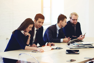 Group of young prosperous designers using mobile phone and discuss ideas in modern interior, successful architects talking among themselves while waiting for the beginning of the business conference
