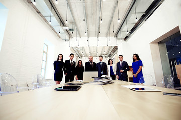 Obraz premium Young creative multi ethnic group of architects posing while standing near table with open laptop computer and paper documents, purposeful business men and women in formal wear resting after briefing