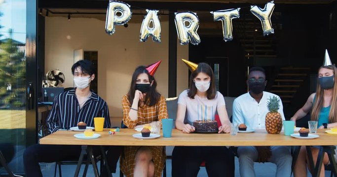 Party after COVID-19. Young upset sad multiethnic friends in masks feeling wrong at birthday celebration slow motion.