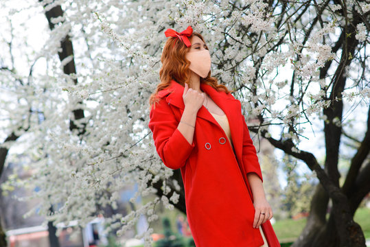 Close up portrait of tender red-haired girl in a red coat under a blossoming cherry tree with a mask with flowers on from the coronavirus.