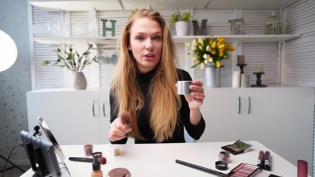 Beauty blogger woman filming daily makeup routine tutorial at camera on tripod. Influencer lady live streaming cosmetics product review in home studio. Vlogger female applies skin powder with brush.