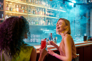 Moments of Happiness. Two women having fun, drinking cocktails while sitting at the bar counter. Friends spending time at night club, restaurant - obrazy, fototapety, plakaty