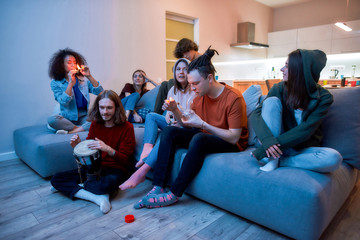 Young guy lighting marijuana in the glass bong and smoking it, relaxing with friends on the sofa at home, young people smoking weed together - obrazy, fototapety, plakaty