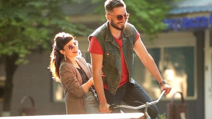 Young couple riding on bicycle together in the park. Romantic boyfriend cycling with happy girlfriend on cargo rack on city street. Woman hugs man having fun riding on bike in tandem. Slow motion. - Powered by Adobe
