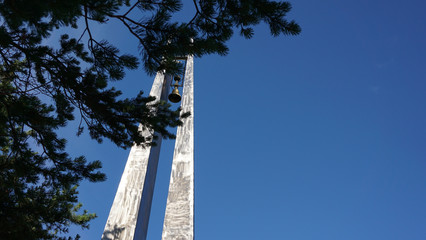 War memorial with a bell against a blue sky. Pine branches