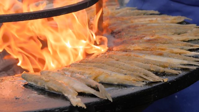 Slow motion: process of cooking breaded european smelt fish on black brazier at summer outdoor food market: close up. Seafood, barbecue, gastronomy, cookery, street food concept