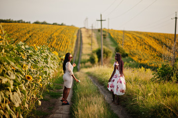 Two pretty young black friends woman wear summer dress pose in a sunflower field.