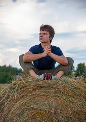 Naklejka na ściany i meble A handsome young man is sitting on a haystack against a blue cloudy sky.