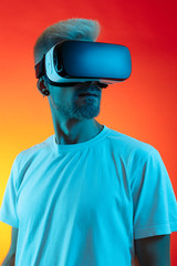 Bearded hipster man with blonde hair wearing virtual reality goggles in studio in neon lights.