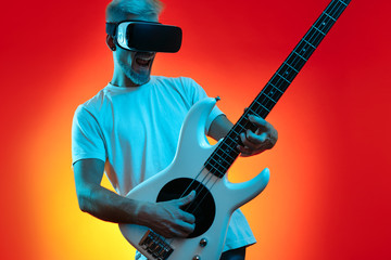 Excited hipster man play white bass guitar with vr headset on the face in neon lights. Music concept. Virtual reality.