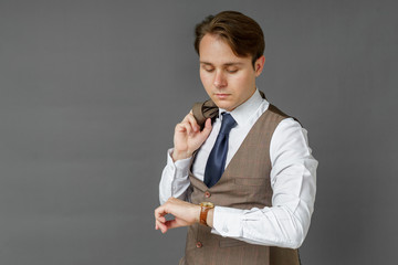 Portrait of a businessman who looks at his watch and holds a jacket over his shoulder. Gray background.