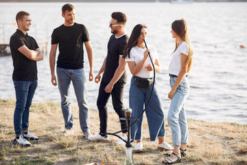 Obraz premium Group of friends is sitting near the lake. They wear casual clothes. Young people eat pizza outside.