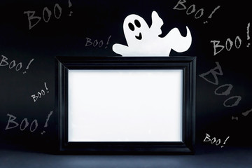 Background for Halloween. Black frame with free space on a black background with lots of BOO! A white ghostly figure peeks out from behind an empty black photo frame. Halloween ideas, concept.