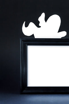 Background for Halloween. Black frame with free space on a black background. A white ghost figure peeks out from behind an empty black photo frame. Halloween typography concept. Halloween Ideas.