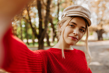 Closeup photo of a charming blonde in red sweater making beautiful selfie in autumn park. Gorgeous...