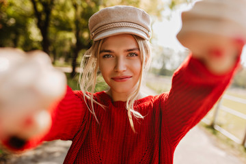 Lovely blonde girl in nice trendy hat and red pullover making selfie in the autumn park.