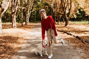 Stunning young woman in trendy red sweater and beige shorts having fun in the beautiful autumn park with her dog.