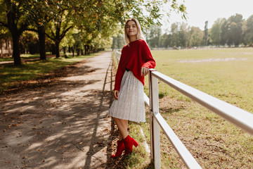 Attractive blonde posing in stylish red outfit in the autumn park. Pretty girl wearing white dress having good time outdoor.