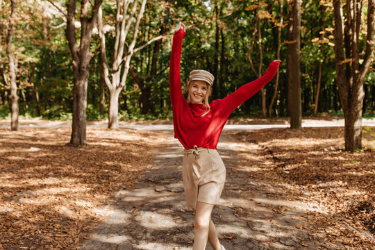 Happy blonde woman in nice red sweater and beige shorts dancing in autumn park. Stylish young woman posing with joy in good weater outdoor.