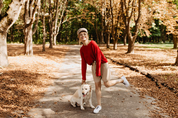Cheerful blonde in stylish red pullover and beige shorts having fun in the beautiful autumn park with her dog.