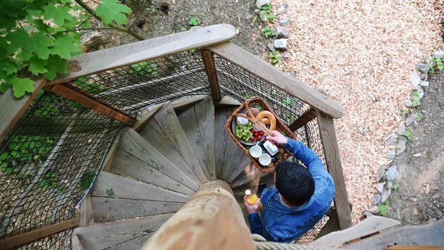 Static Overhead Slow Motion FHD Shot Of A Man Wearing Jeans And A Denim Jacket Climbing A Wooden Spiral Staircase Of A Treehouse, Carrying A Basket With Breakfast. Man Walking Up The Stairs With Food.