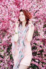 A young beautiful fashionable girl in a stylish spring dress and shawl with floral patterns. The smiling redhaired model poses outside in blossoming pink trees. Spring fashion concept