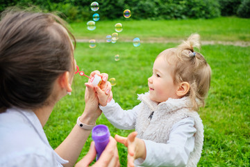 Happy toddler and mom make soap bubbles in a summer park.