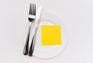 A big white plate, knife and fork lie on a tablecloth. Empty sheet of yellow paper for copy space and mockup, top view.