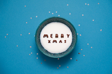 Top view of bowl with milk and chocolate alphabet cereals. Merry xmas text. Blue background
