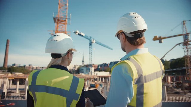 Male Civil Engineer and Young Female Building Architect Use a Tablet Computer on a City Construction Site. They Talk About the Future of Real Estate Development and Planning. Wearing Safety Hard Hats.