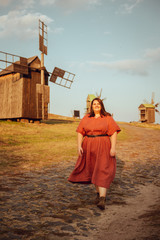 Beautiful and confident plus size model in red dress and brown boots  posing outdoors at autumn landscape with wooden mill at background