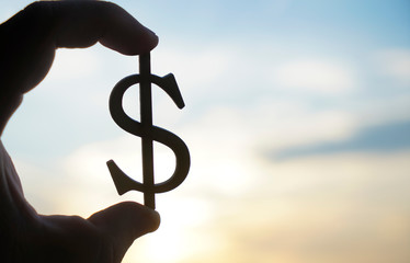 Hand hold silhouette of dollar sign , symbol of american money against sun rays and sunset sky with...