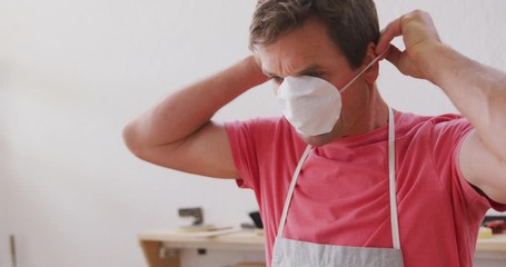 Caucasian male surfboard maker wearing a protective apron and putting on a face mask - Powered by Adobe