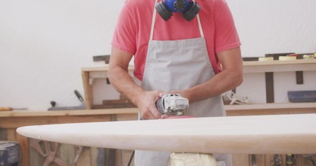 Caucasian male surfboard maker wearing a face mask and preparing to polishing a surfboard - Powered by Adobe